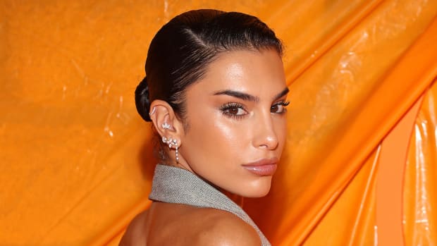 Jessica Aidi wears a slicked-back bun and a gray halter top and poses in front of an orange backdrop.